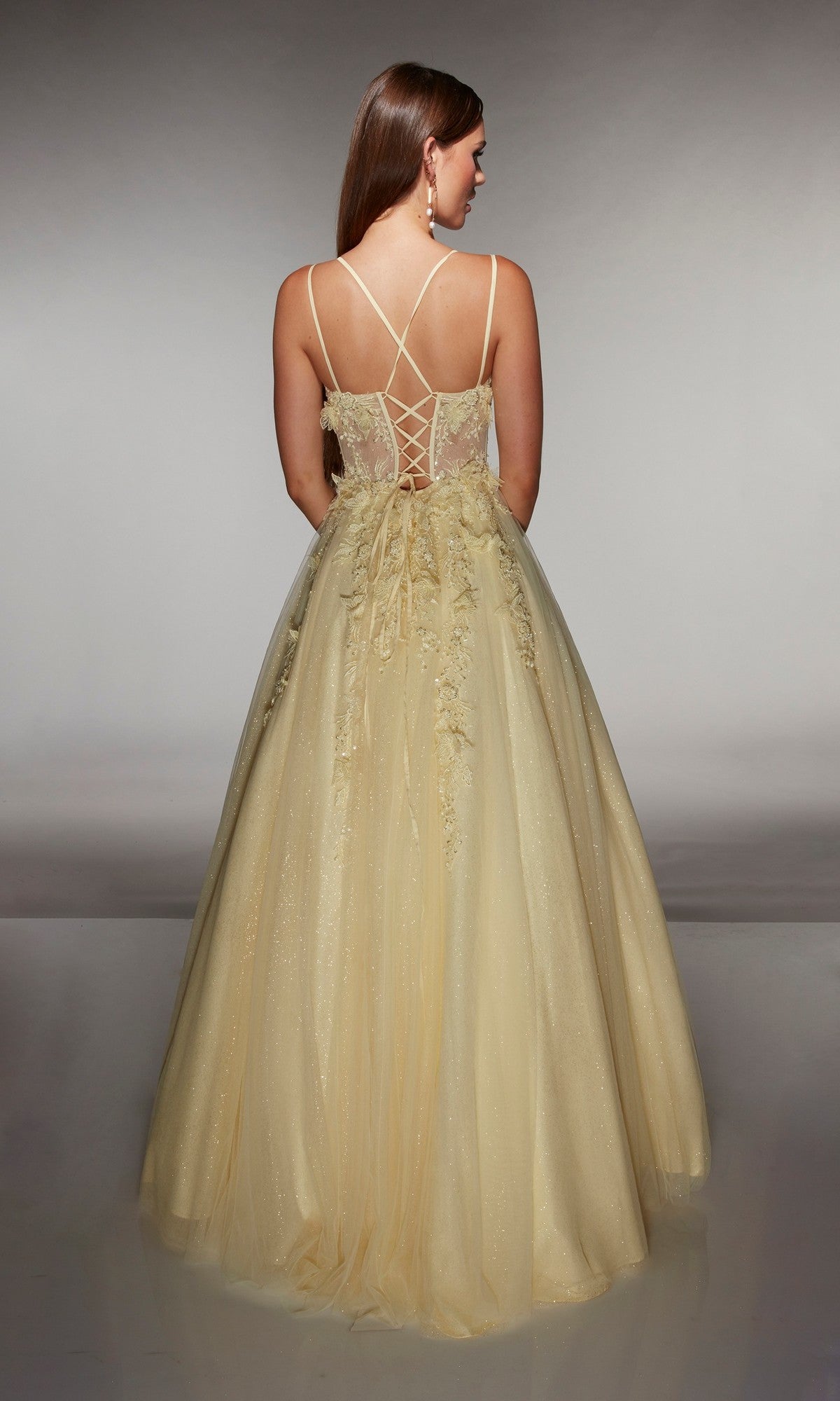 A woman models the Alyce 62203 long prom dress, featuring a floor-length yellow design with a lace-up back, floral appliqué accents, and glitter tulle for a subtle shimmer, all set against a gray background.